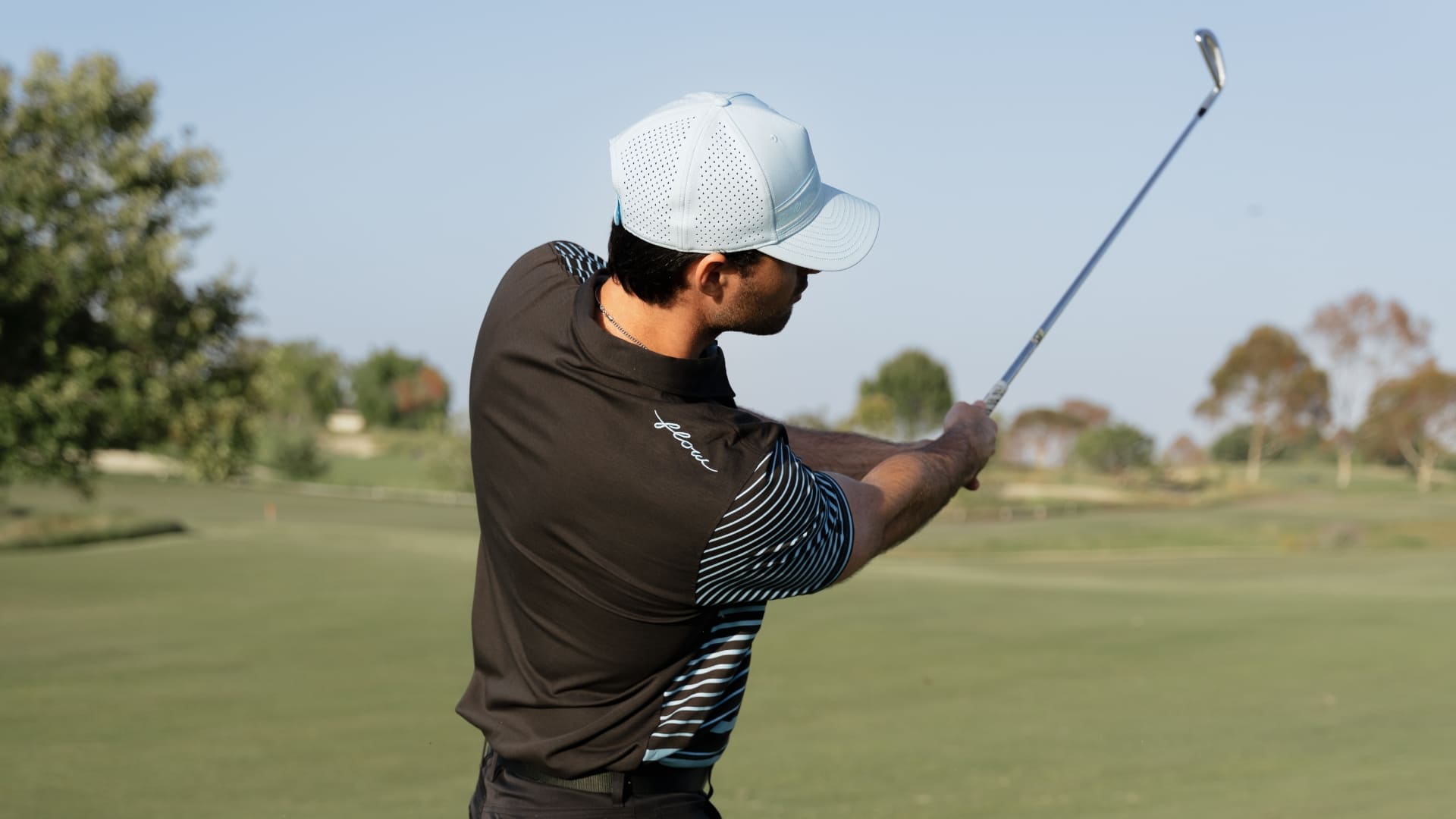 A male golfer in a black polo shirt with striped sleeves and a light blue cap, captured mid-swing on a golf course.